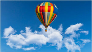 Ein Heißluftballon mit farbenfroher Ballonhülle schwebt vor weißen Wolken am blauen Himmel. Im Fahrkorb sind die Passagiere zu sehen.