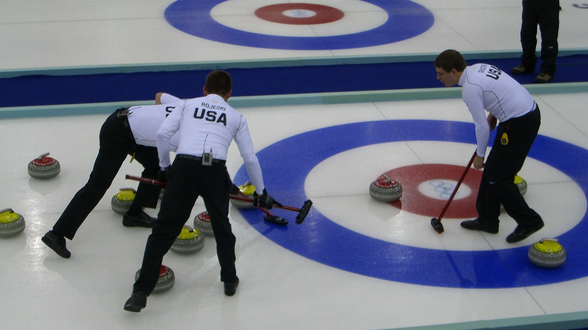 Drei Curling-Spieler stehen auf dem Eis beim Zielpunkt. Sie wischen mit ihren Besen die Eisbahn, damit ihr Curling-Stein an den anderen Steinen vorbei näher an das Ziel gelangt.