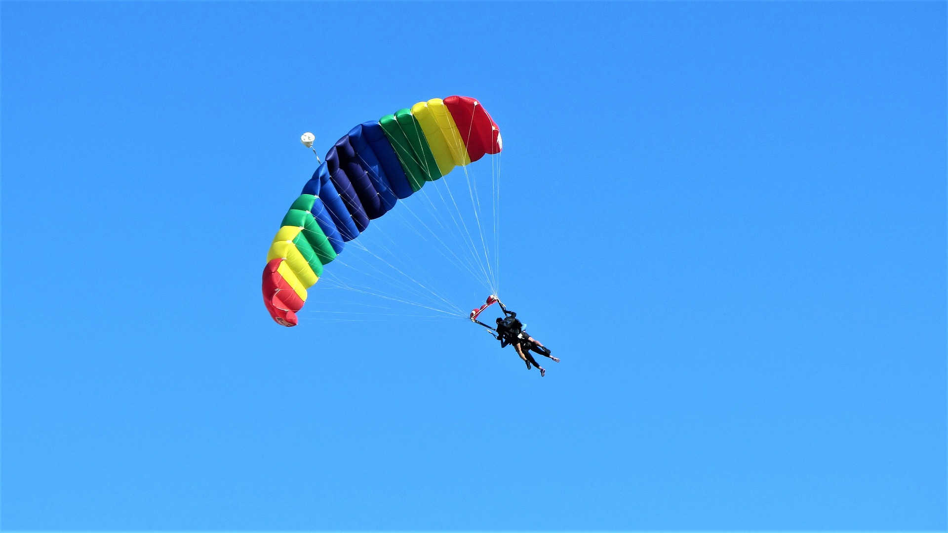 Auf diesem Bild ist ein Tandem-Fallschirmsprung zu sehen. Zwei Personen schweben an einem bunt gestreiften Fallschirm dem Erdboden entgegen.