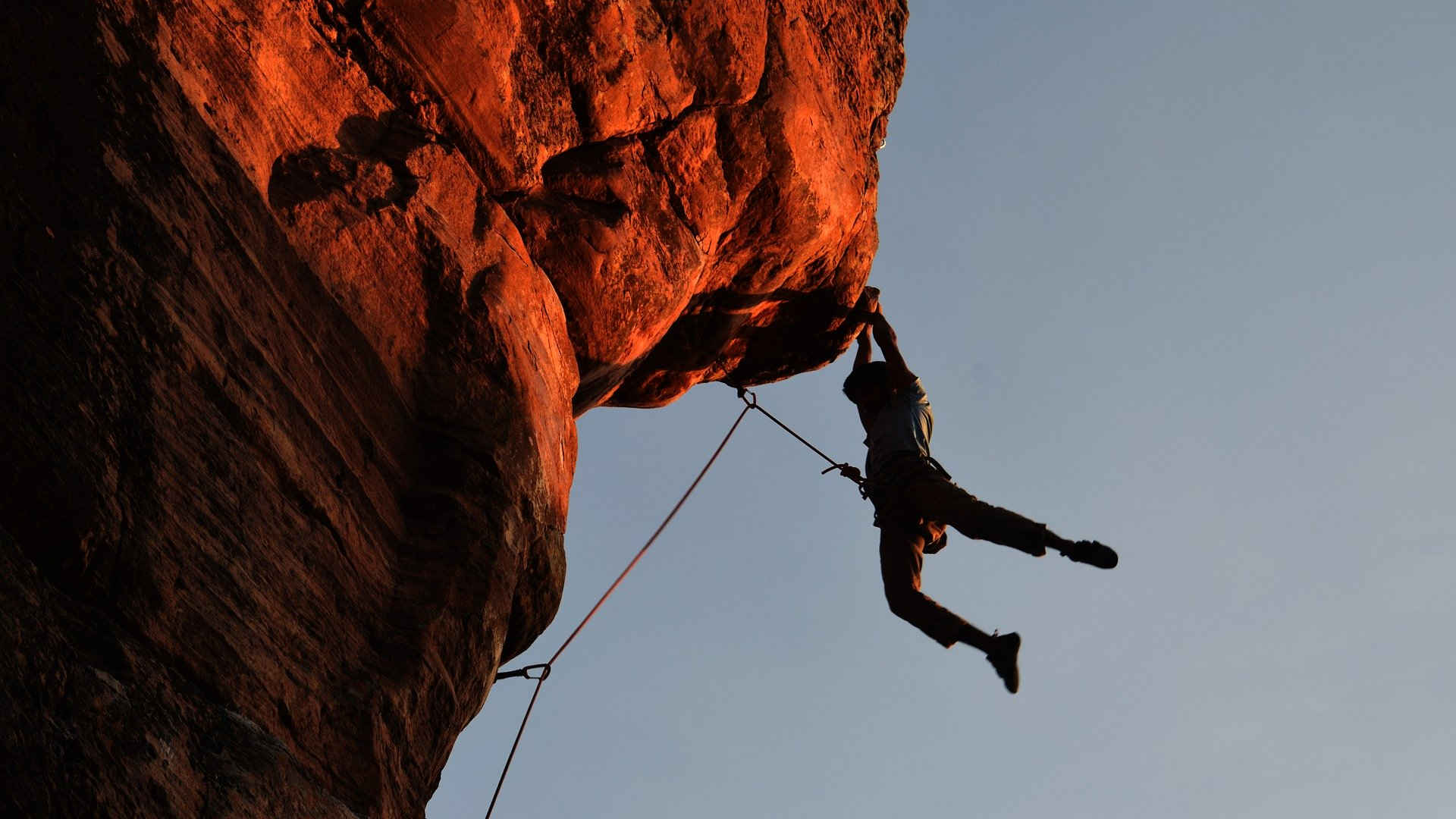 Wir sehen einen Freikletterer, der sich am Fels an einem Überhang hoch arbeitet. Er ist mit einem Seil gesichert. Er hat nur Halt mit den Händen, die Beine hängen frei in der Luft.