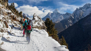 Wanderer gehen auf einem schneebedeckten Weg. Sie tragen große schwere Rucksäcke mit Gepäck für eine längere Wanderung. Im Hintergrund sehen wir schneebedeckte Berge vor blauem Himmel mit weißen Wolken.