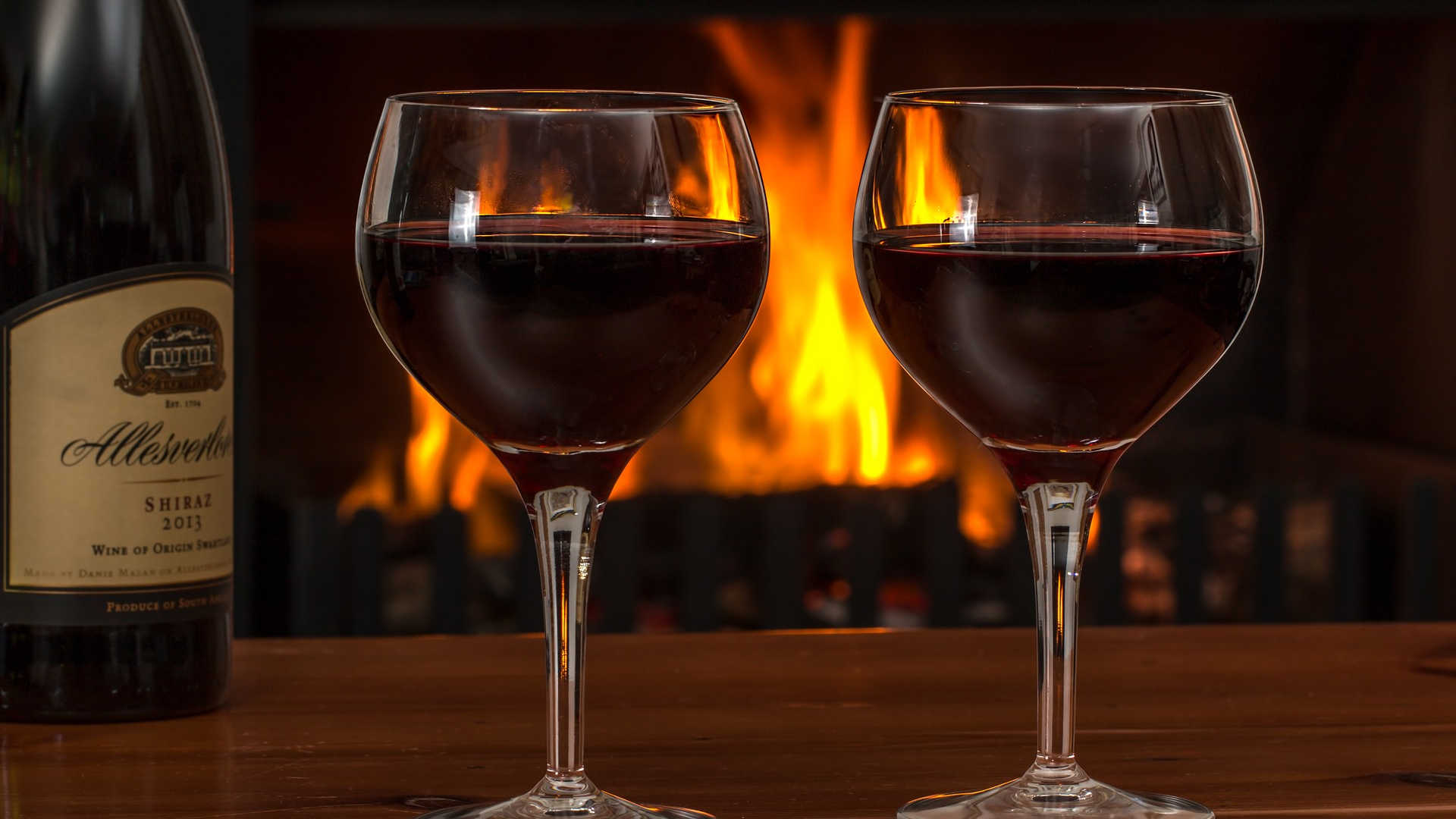 Es stehen zwei mit Rotwein gefüllte Gläser einladend auf einem Tisch. Im Hintergrund lodert das gemütliche Kaminfeuer.