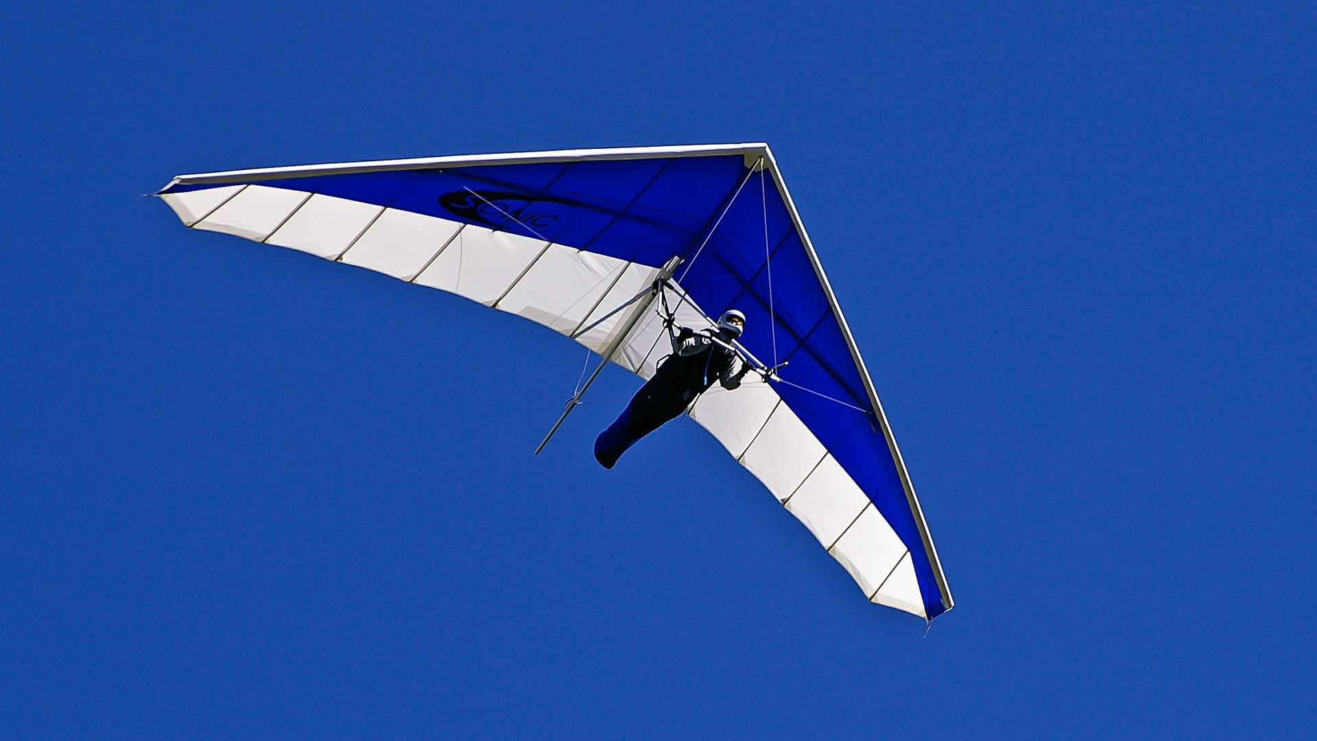 Wir sehen einen klassischen Drachen in blau-weiß. Der Pilot hängt liegend in seinem Gurtzeug, welches ihn ähnlich wie ein Schlafsack umschließt. Er steuert seinen Hanggleiter durch Gewichtsverlagerung.