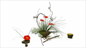 In der Mitte steht eine Keramikschale mit Blumen und einem Zweig. Links davor ziert eine Blüte einen Stein. Ein trockener Zweig und eine kleine Schale ergänzen das Ikebana-Arrangement.