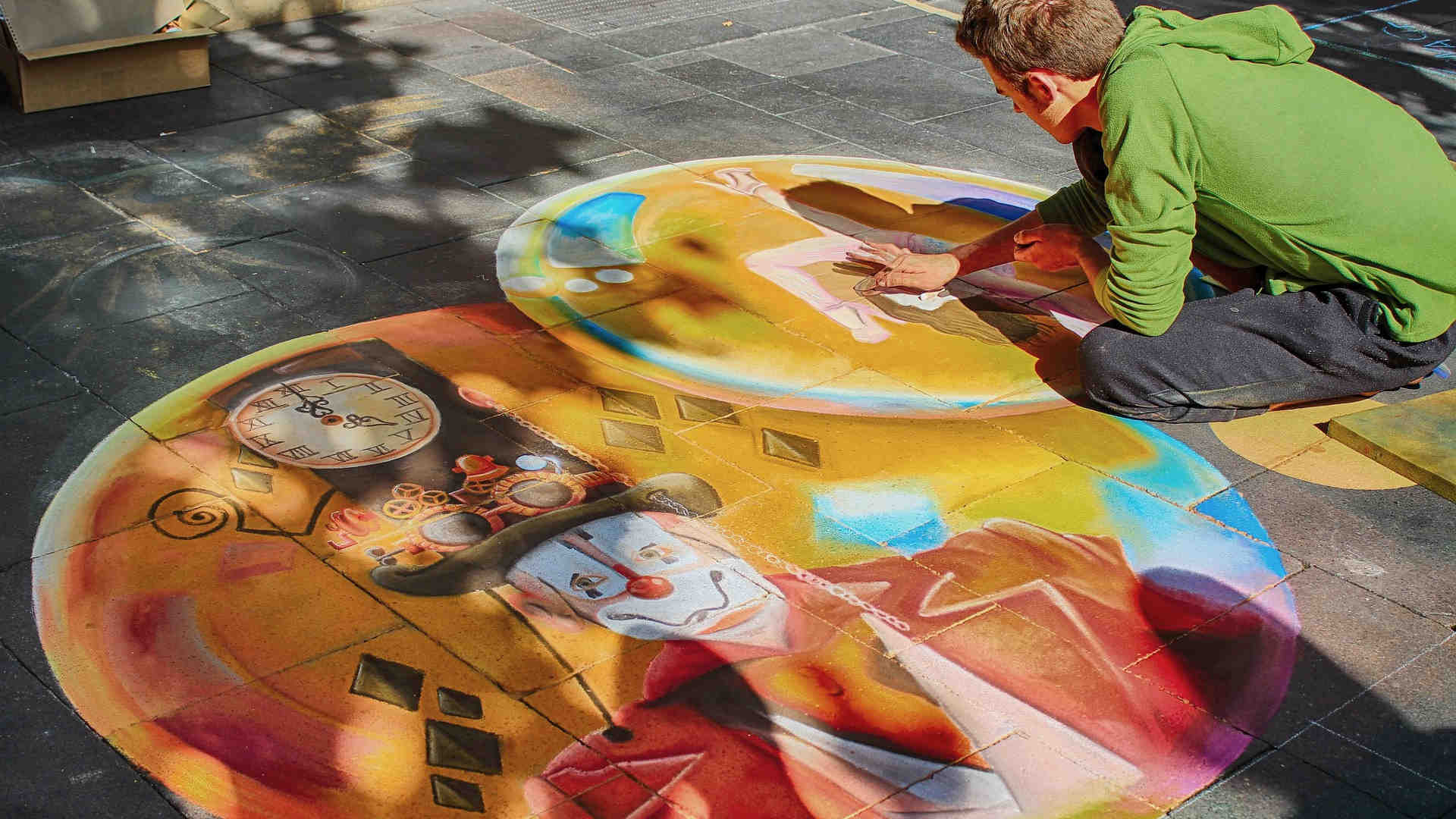 Wir sehen einen Straßenmaler, wie er ein Bild aus seiner Fantasie auf die Platten der Fußgängerzone malt. Ein Teil davon ist schon fertig und zeigt einen Clown.