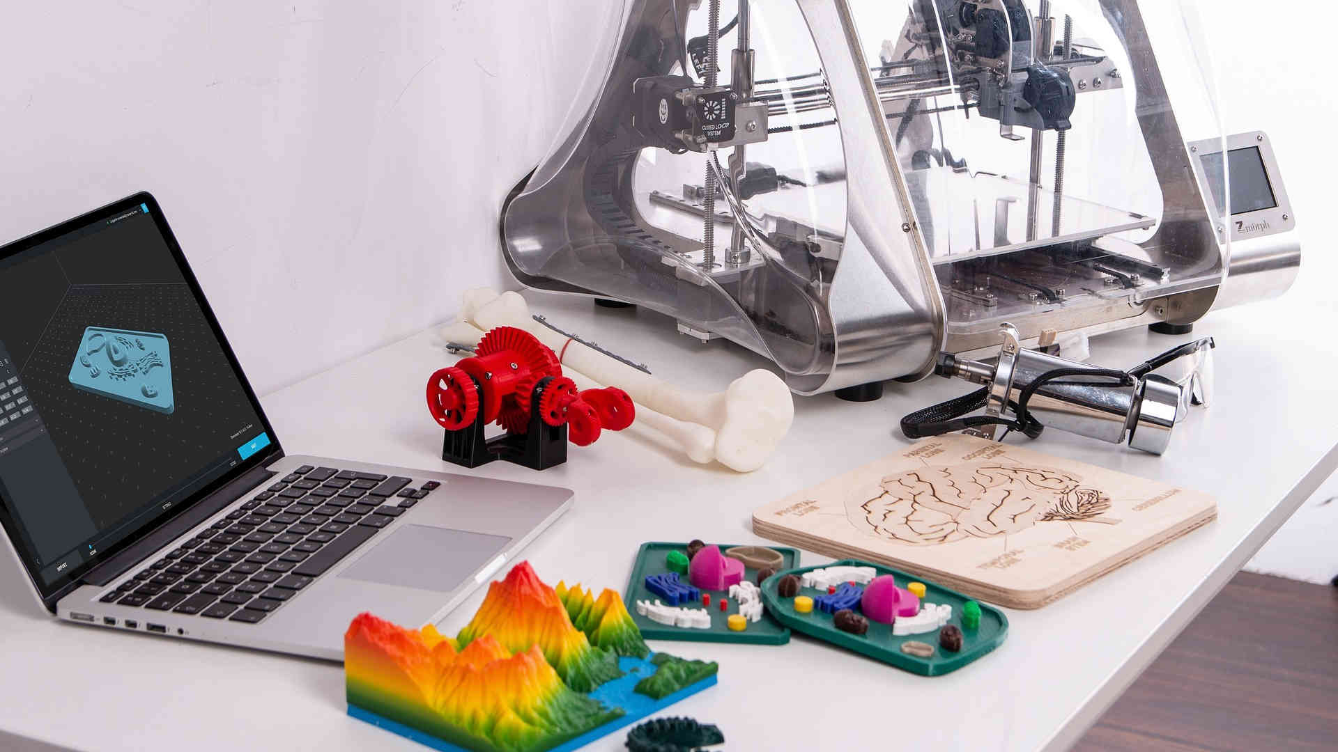 Auf dem Tisch stehen ein 3D-Drucker und ein Laptop mit einer 3D-Software. Davor liegen einige bunte Gegenstände, die schon mit dem Drucker hergestellt wurden.
