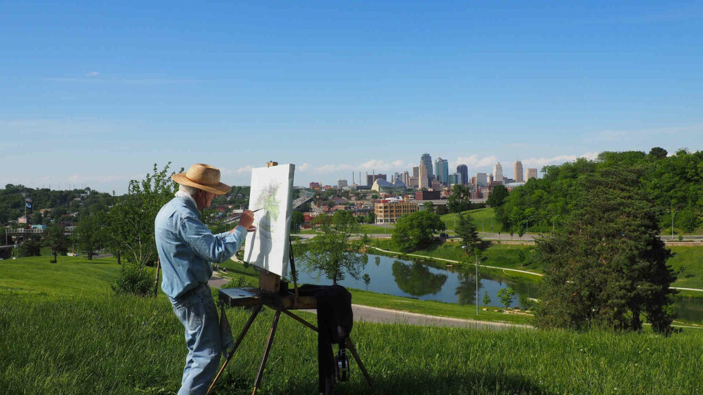 Hobby finden: Malen - Mit Blick auf die Natur und eine Skyline im Hintergrund steht ein älterer Herr an seiner Staffelei. Er bringt dieses Motiv ganz in sein Tun versunken auf die Leinwand.