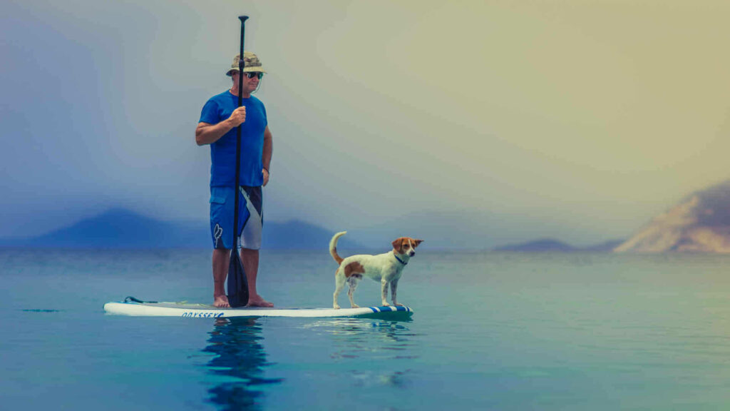 Hobby finden: Stand Up Paddling - Der Mann mit dem Paddel in der Hand lässt sich mit seinem Board langsam über das Wasser treiben. Sein Hund steht an der Spitze des Boards.
